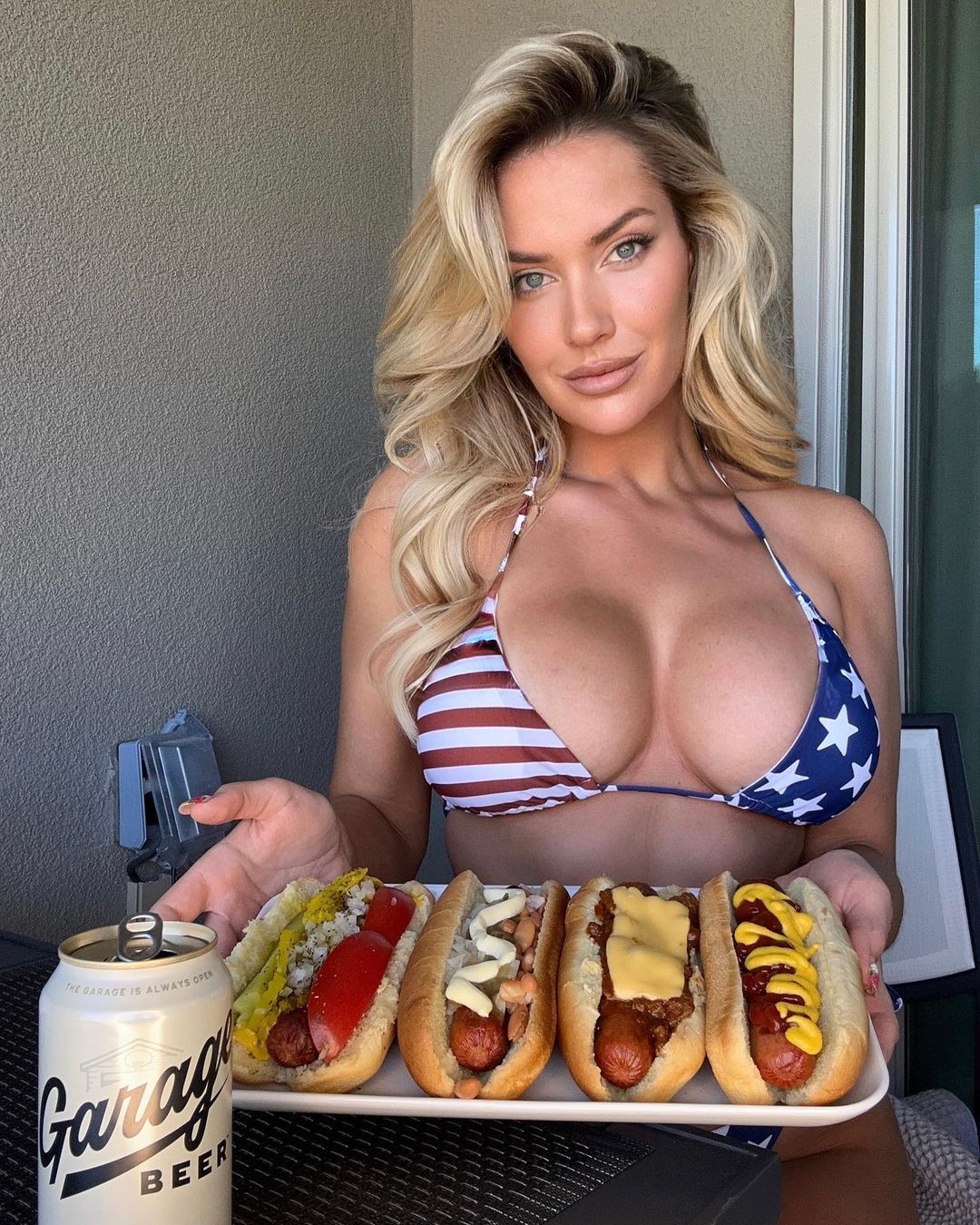 a woman in a bikini is holding a tray of hot dogs and a can of garage beer