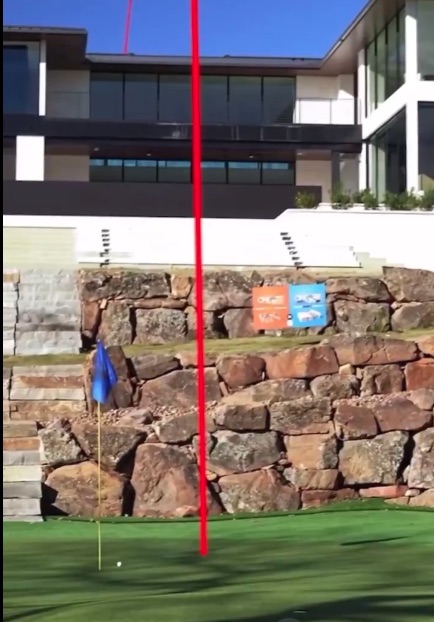 Putting green with a golf ball near a flag, behind a rock wall and modern house.