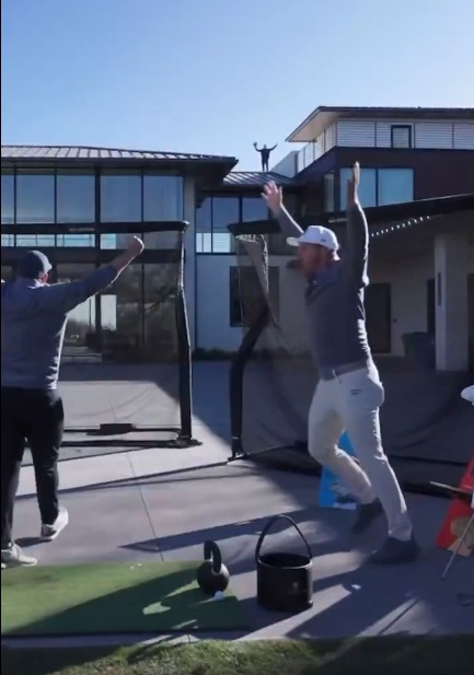 Two men celebrating on a golf course, one jumping for joy while the other raises his arms. A man is seen standing on a house roof in the background.