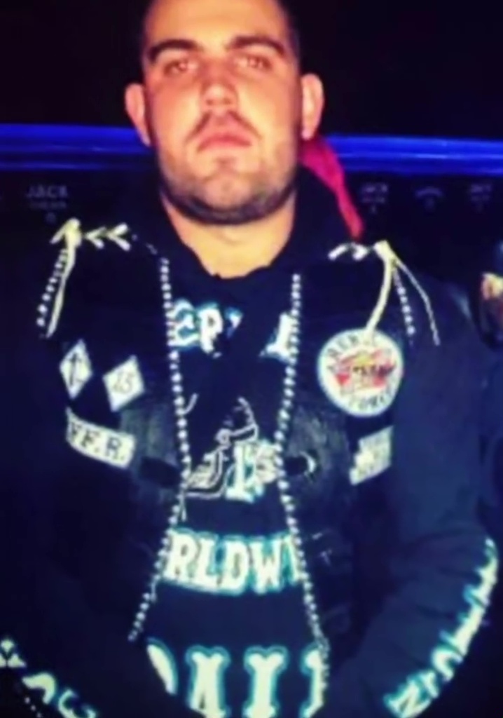 Photo of a man wearing a dark blue hoodie with patches and a beaded necklace.