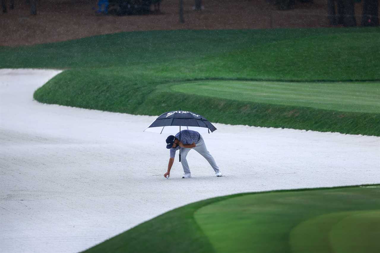 Golfer under umbrella placing ball in sand trap.