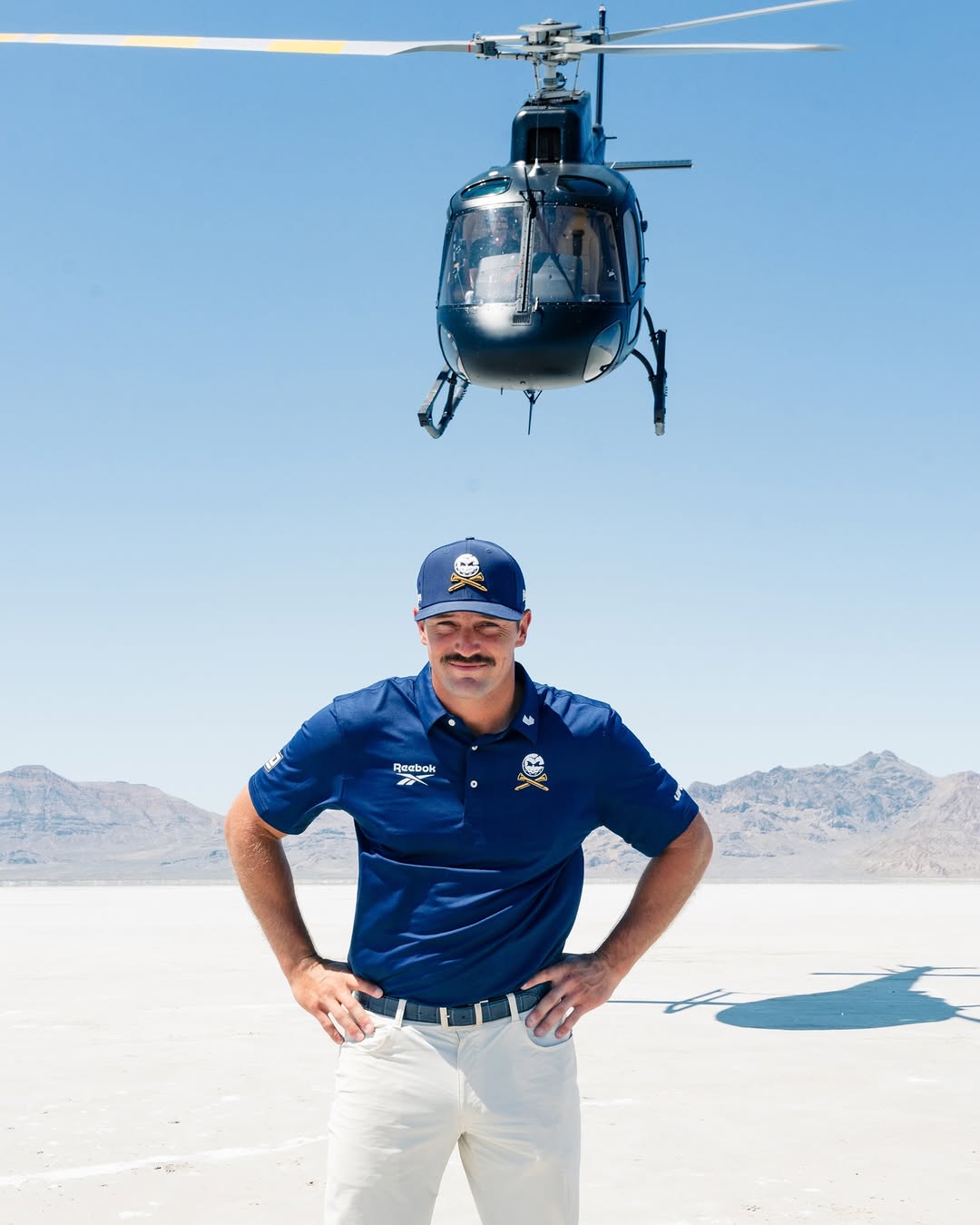 Bryson DeChambeau standing in a desert with a helicopter overhead.