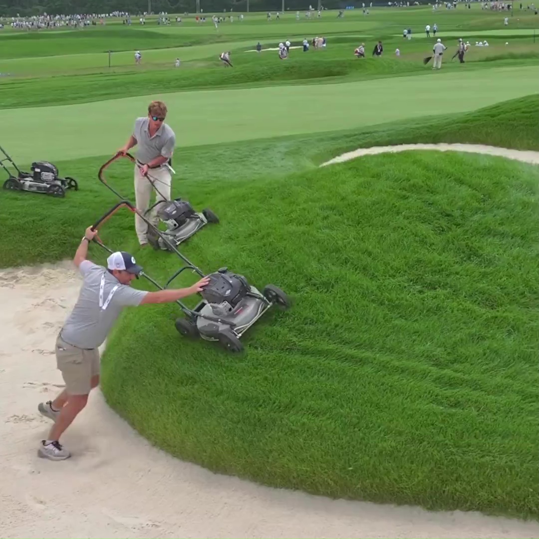 Two groundskeepers using lawnmowers on a golf course.