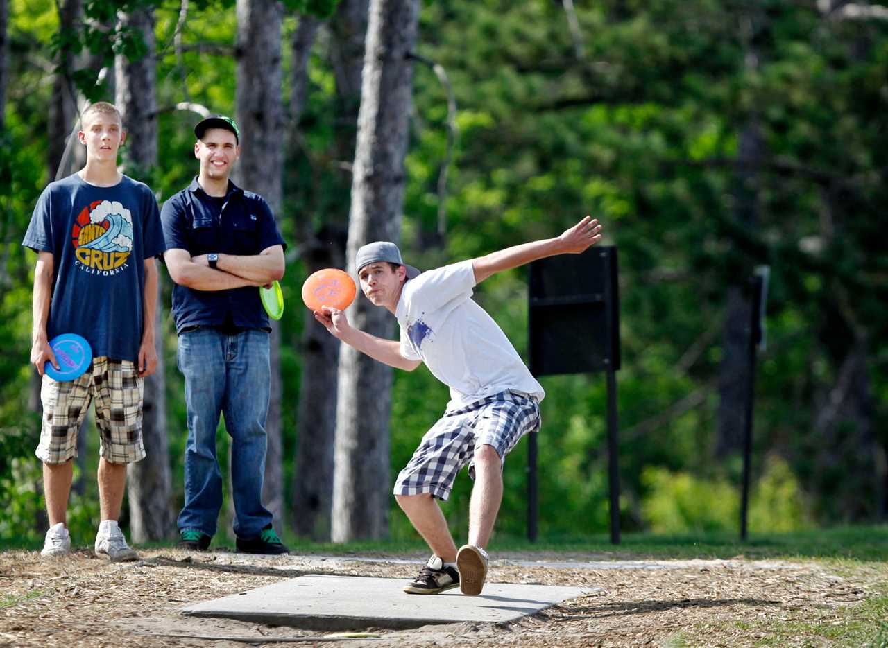 Man throwing a disc at a disc golf course while two other men watch.