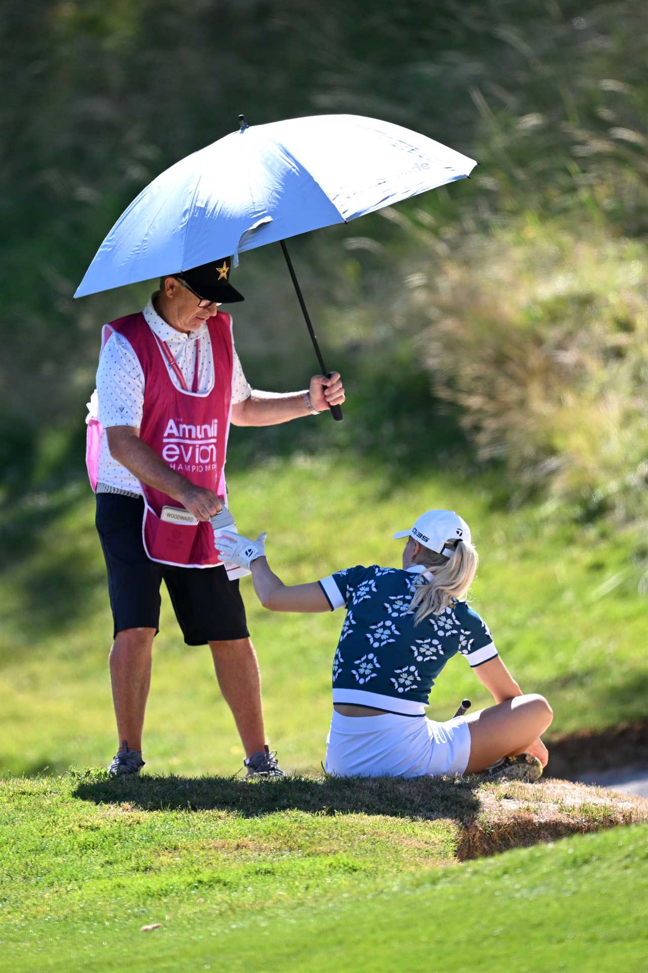 A caddie holding an umbrella over a seated golfer and handing her a drink.