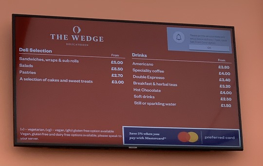 Menu board for The Wedge Deli showing prices for sandwiches, salads, pastries, cakes, and drinks.