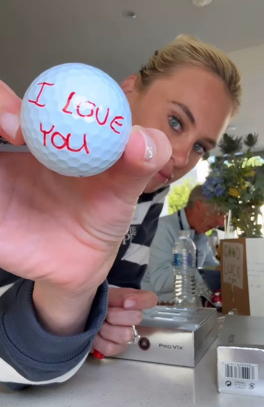 Close-up of a hand holding a golf ball with "I love you" written on it.